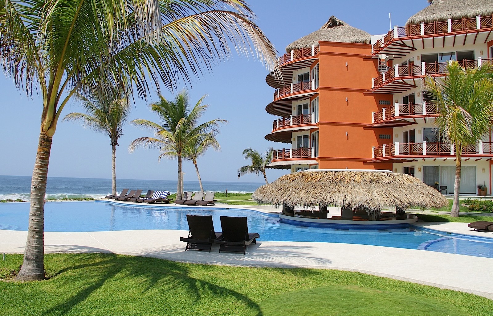 Resort pool area with palm trees, lounge chairs, a thatched-roof bar, and a multi-story orange building beside it