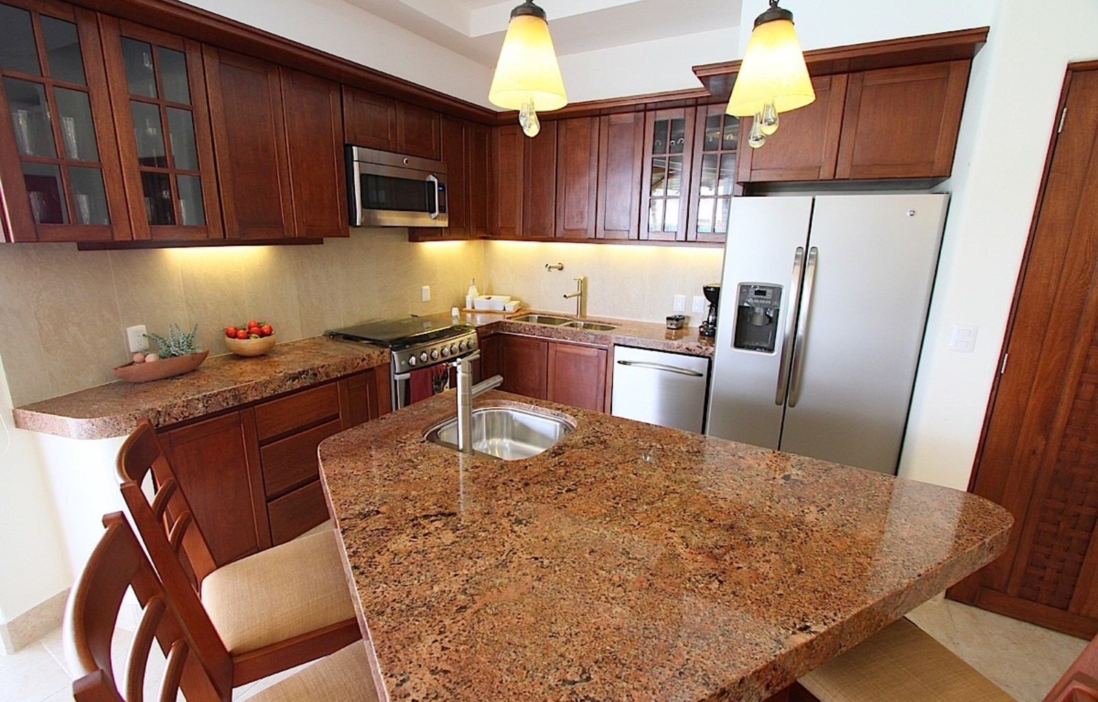 Modern kitchen interior with dark wood cabinetry, granite countertops, stainless steel refrigerator, and pendant lighting