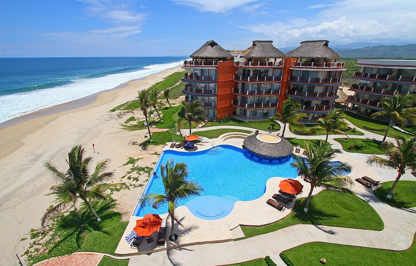 Aerial view of a beachfront resort complex with curved swimming pool, orange umbrellas, and thatched palapa structures
