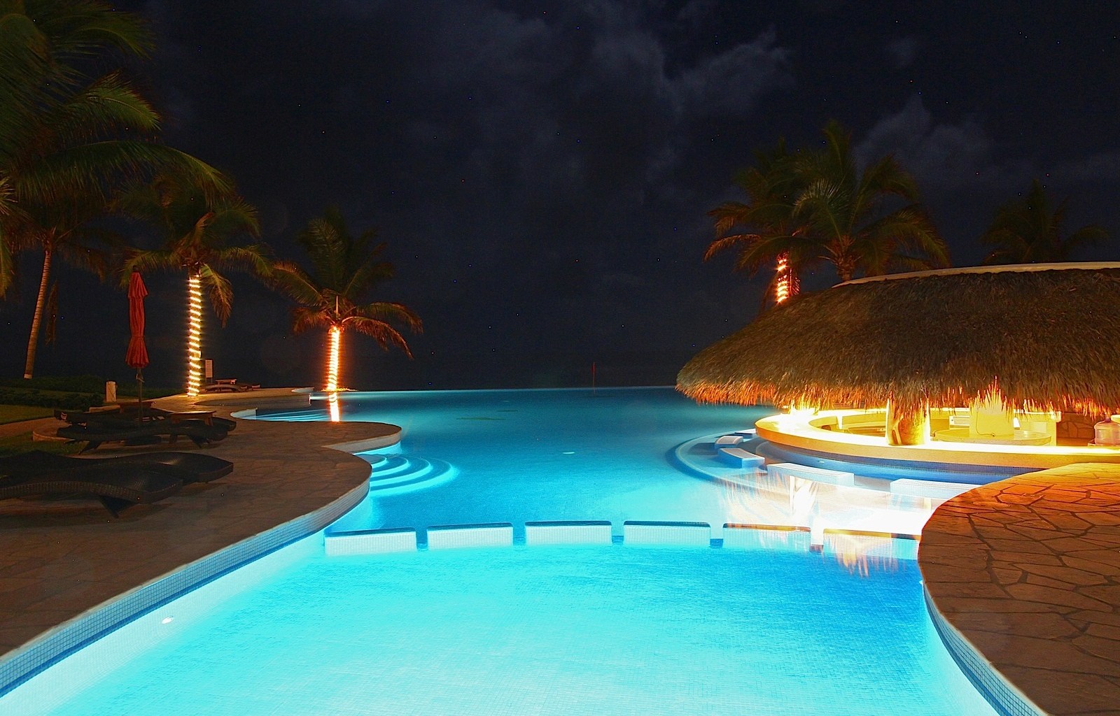 Illuminated infinity pool at night with thatched-roof bar, palm trees, and lounge chairs glowing blue and orange