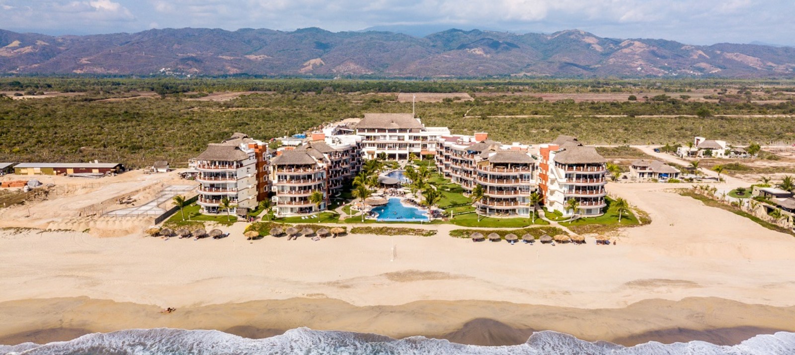 Aerial view of a beachfront resort complex with multiple buildings, pool, and surrounding countryside and mountains