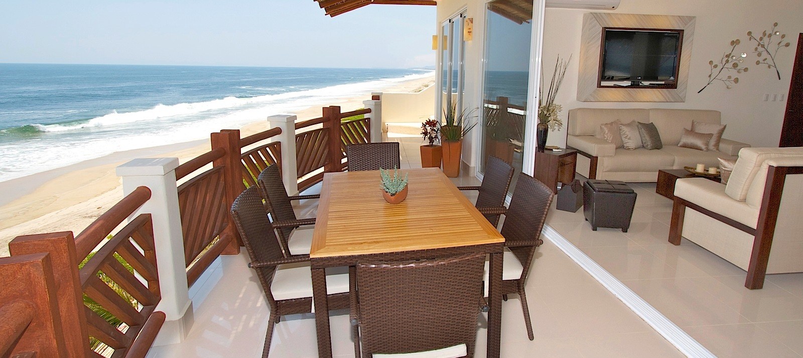 Oceanfront balcony with wooden dining table and chairs, living room visible indoors, wide sea view beyond the railing